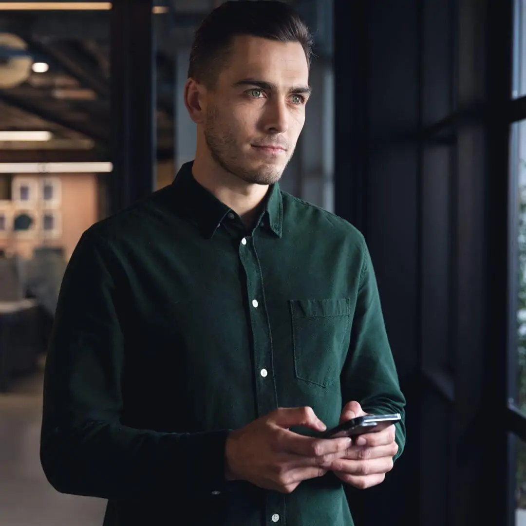 Homem em ambiente corporativo segurando um smartphone, olhando pela janela. Ilustra a seção sobre a transformação de ativos e direitos creditórios em liquidez pela Value Securitizadora.